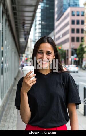 Porträt der jungen Frau, die mit Papierbecher in der Hand breit lächelte. Urbaner Hintergrund. Mittlere Aufnahme. Stockfoto