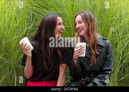 Zwei junge Frauen, die einmal-Tassen halten, die sich mit einem breiten Lächeln betrachten. Grashintergrund. Stockfoto