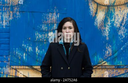 Attraktive junge weiße Frau mit braunen Haaren, die gegen eine blaue Graffitiwand posiert Stockfoto
