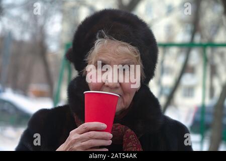 Seniorinnen tragen warme Kleidung, die roten Plastikbecher in einer Hand hält, Außenporträt Stockfoto