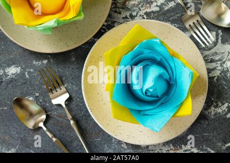 Dekorative Blume aus farbigen Papierservietten. Tischeinstellung mit farbigen Papierservietten auf Steingrund, flacher Lay. Stockfoto