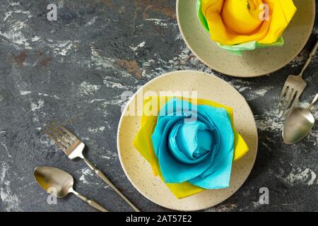 Dekorative Blume aus farbigen Papierservietten. Tischeinstellung mit farbigen Papierservietten auf Steingrund, flacher Lay. Freier Speicherplatz für Ihren Text. Stockfoto