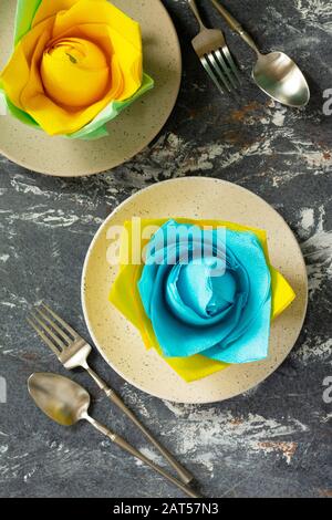 Dekorative Blume aus farbigen Papierservietten. Tischeinstellung mit farbigen Papierservietten auf Steingrund, flacher Lay. Stockfoto