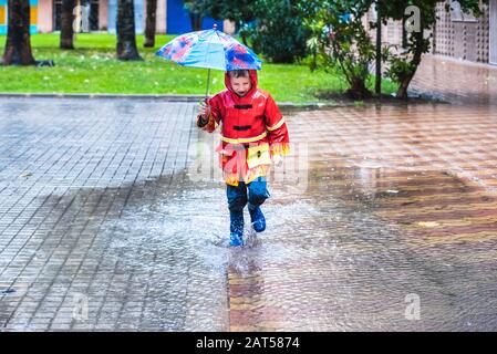 Junge mit rotem Regenmantel, verkleidet mit Regenschirmspritzern in einer Pfütze Wasser. Stockfoto