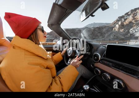 Fröhliche Frau in hellem Hut und Jacke, die mit einem Cabrio-Auto unterwegs ist, während sie bei Sonnenuntergang auf der Wüstenstraße unterwegs ist. Sorgloses Lifestyle- und Reisekonzept Stockfoto