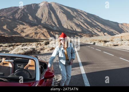 Junge Frau, die auf der Straße im Wüstental unterwegs ist und in der Nähe des Cabrio-Wagens am Straßenrand steht, mit Vulkan im Hintergrund Stockfoto