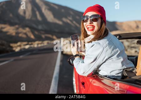 Frau, die mit dem Auto auf dem vulkanischen Tal unterwegs ist, sich aus dem Autofenster lehnt und mit einem Smartphone fotografiert Stockfoto