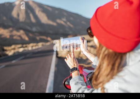 Frau, die mit dem Auto auf dem vulkanischen Tal unterwegs ist, sich aus dem Autofenster lehnt und mit einem Smartphone fotografiert Stockfoto