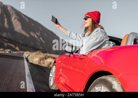 Frau, die mit dem Auto auf dem vulkanischen Tal unterwegs ist, sich aus dem Autofenster lehnt und mit einem Smartphone fotografiert Stockfoto