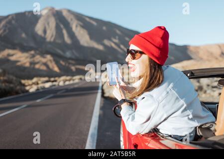 Glückliche Frau, die mit dem Auto auf dem vulkanischen Tal unterwegs ist, sich aus dem Autofenster lehnt und Smartphone mit SMS zum Reisen hält Stockfoto