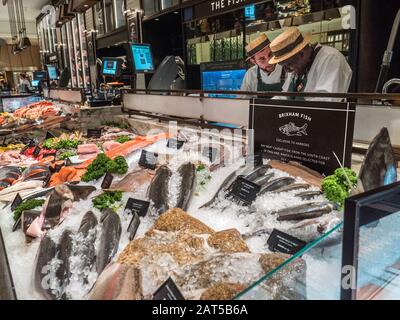 Harrods Food Hall mit "The Fishmonger" mit einer großen Auswahl an exklusiven handverlesenen Brixham UK Fisch Fisch Meeresfrüchte Crustacea zum Verkauf, Verkaufsassistenten mit traditionellen Strohboater-Hüten, Vorbereitung einer Kundenauswahl, Harrods Food Hall Fischhändler Kaufhaus Mall Shop, Brompton Rd, Knightsbridge, London SW1X 7XL Stockfoto