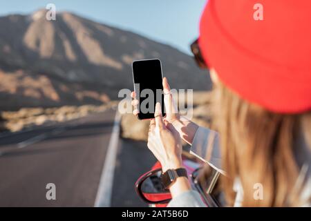 Frau, die mit dem Auto auf dem vulkanischen Tal unterwegs ist, sich aus dem Autofenster lehnt und mit einem Smartphone fotografiert. Telefon mit leerem Bildschirm zum Kopieren des Einfügevorgangs Stockfoto