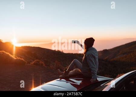 Junge Frau fotografiert mit Telefon schöne Landschaft während eines Sonnenuntergangs und sitzt auf der Autohaube, während sie hoch in den Bergen unterwegs ist Stockfoto