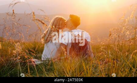 Liebevolles Paar sitzt und küsst auf einer Bergwiese und genießt den Blick auf den Sonnenuntergang. Ein Berghügel. Wundervolle Landschaft der Berge. Verliebt. Enj Stockfoto