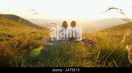 Liebevolles Paar sitzt und küsst auf einer Bergwiese und genießt den Blick auf den Sonnenuntergang. Ein Berghügel. Wundervolle Landschaft der Berge. Verliebt. Enj Stockfoto