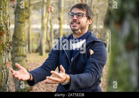 Fröhlicher junger Mann verspielte Wurfblätter mit offenen Armen, die unter einem Regen von Blättern im Herbstpark tanzen. Stockfoto