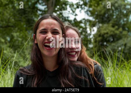 Zwei Freundinnen verschiedener Rassen lachen mit einer Umarmung der anderen von hinten. Grüner Parkhintergrund. Stockfoto