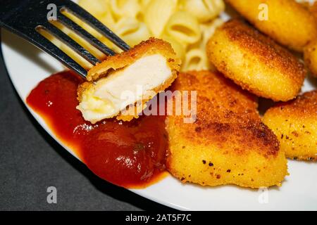 Ein Stück Nuggets auf einer Gabel und ein weißer Teller mit gebratenen Hühnerhuhn Nuggets, Pasta und Ketchup auf dunklem Hintergrund. Nahaufnahme Stockfoto