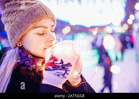 Wunderschönes Mädchen trinkt im Winter heißen Tee aus Pappglas auf dem Hintergrund der abendlichen Bokeh-Beleuchtung Stockfoto