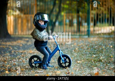 Ein Junge mit einem Motorradhelm für Erwachsene am Kopf und einem Roller im Park Stockfoto