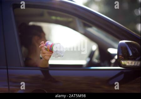 Eine junge Frau wirft Müll aus dem Auto - eine Kaffeetasse und eine Tasche. Umweltverschmutzung, selektiver Fokus, Hintergrundbeleuchtung. Kopierbereich Stockfoto