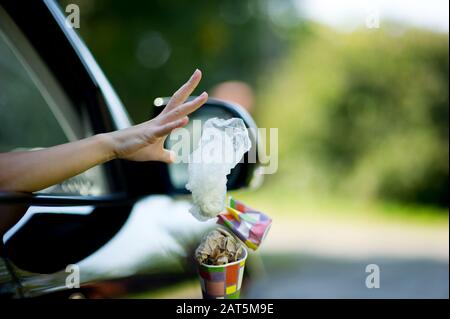 Eine junge Frau wirft Müll aus dem Auto - eine Kaffeetasse und eine Tasche. Umweltverschmutzung, selektiver Fokus, Hintergrundbeleuchtung. Kopierbereich Stockfoto