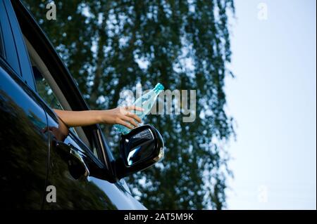 Eine junge Frau ist dabei, eine leere Plastikflasche in ein offenes Autofenster zu werfen. Ansicht von unten, auf dem Hintergrund verschwommener Bäume und Himmel. Kopieren Sie Spac Stockfoto