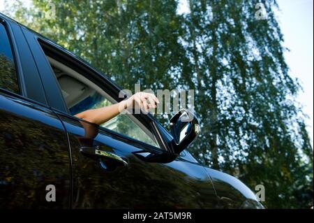 Eine junge Frau hält einen entspannten Arm außerhalb des Autos - sie hält ihre Hand durch das offene Fenster des Vordersitzes eines dunklen Autos heraus. Ansicht von unten, auf Stockfoto