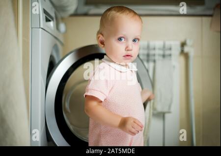 Blauäugiges Mädchen mit rosafarbenen Overalls steht in der Nähe der Waschmaschine und blickt auf die Kamera. Stockfoto