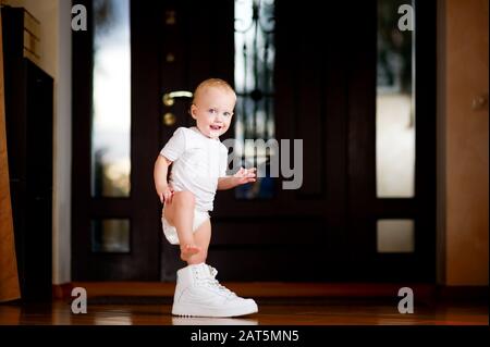 Kleines Mädchen, das versucht, einen großen weißen Sneaker eines Erwachsenen anzuziehen, der im Haus steht Stockfoto