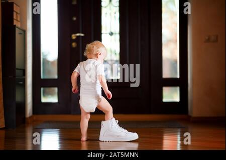 Baby-Mädchen in einem weißen T-Shirt und Windel legt einen weißen Papa Sneaker an, der zu Hause im Flur steht Stockfoto