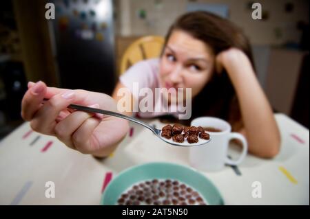 Eine junge Frau zeigt einen Löffel mit Schokoladenkugeln und äußert ihre Zurückhaltung, sie zu essen. Schokoladengetreide mit Milch im Fokus und Mädchen mit Grimace in Stockfoto