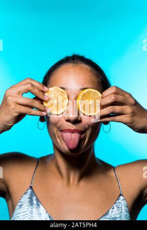 Junge Frau bedeckt Augen mit Zitronenscheiben und sticht Zunge heraus. Blauer Hintergrund. Nahaufnahme. Stockfoto