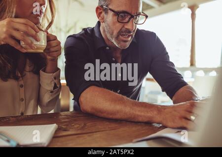 Mann, der sich einen Laptop ansieht und mit einer Kollegin über die Arbeit spricht, die Kaffee hat. Geschäftsleute diskutieren im Coffeeshop über ein neues Projekt über Laptops. Stockfoto