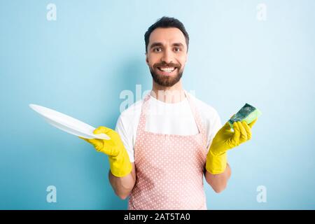 Fröhlicher Mann in gestrichelter Schürze mit Halteplatte und Schwamm auf Blau Stockfoto