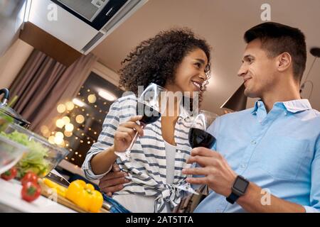 Romantisches Datum. Junges multiethnisches Paar steht beim Küchen-Kochen am Dinner und umarmt Trinkwein, der sich gegenseitig lachend freudige Nahaufnahme ansieht Stockfoto