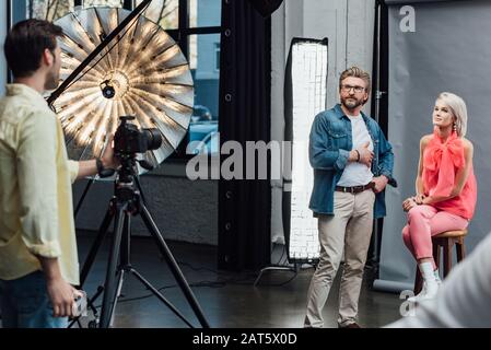 Selektive Fokussierung des gutaussehenden Kunstdirektors, der mit der Hand in der Tasche in der Nähe eines attraktiven Modells steht und den Fotografen betrachtet Stockfoto