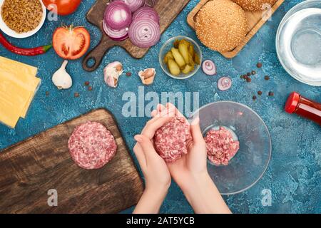 Draufsicht über Frauen, die Schnutschen für frische Burger in der Nähe von Zutaten auf blauer, strukturierter Oberfläche machen Stockfoto