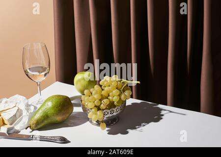 Klassisches Stillleben mit Camembert-Käse, Weißwein und Obst auf dem Tisch in der Nähe von Vorhang isoliert auf Beige Stockfoto