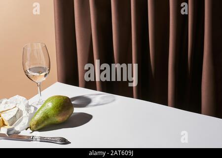 Klassisches Stillleben mit Camembert-Käse, Weißwein und Birne auf dem Tisch in der Nähe von Vorhang isoliert auf Beige Stockfoto