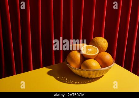 Klassisches Stillleben mit Orangen im Korbkorb auf gelbem Tisch in der Nähe des roten Vorhanges Stockfoto