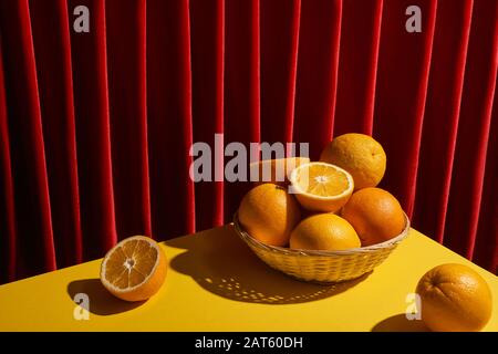 Klassisches Stillleben mit Orangen im Korbkorb auf gelbem Tisch in der Nähe des roten Vorhanges Stockfoto