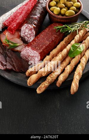 Leckere Fleischplatte mit Oliven und Grissini an Bord auf schwarzer Oberfläche Stockfoto
