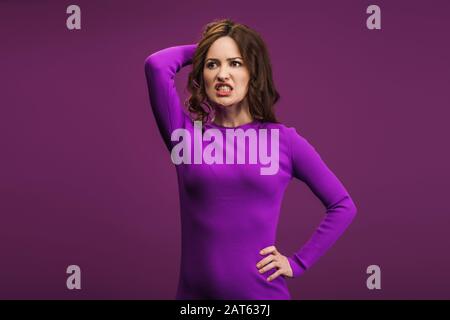 Wütende Frau, die mit der Hand auf der Hüfte steht und den Kopf auf violettem Hintergrund berührt Stockfoto