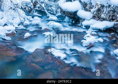 Winterlandschaft eines kleinen Gebirgsflusses mit bunten Steinen, tiefgefrorener Wasserfall in den Alpen. Bäume im Wald und Steine im Bach sind schneebedeckt Stockfoto