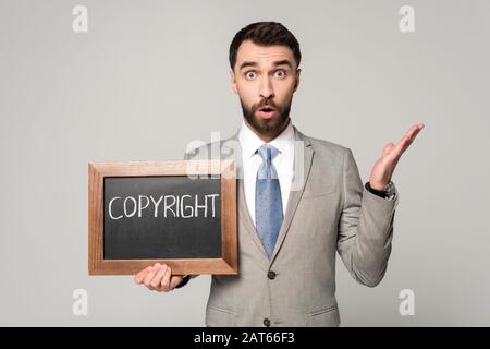 Schockierter Geschäftsmann, der die Kamera betrachtet, während er ein schwarzes Brett mit einer auf Grau isolierten Copyright-Beschriftung hält Stockfoto
