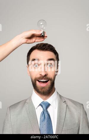 3/4-Ansicht der Frau, die die Glühbirne über dem Kopf des aufgeregten Geschäftsmannes hält, isoliert auf Grau Stockfoto