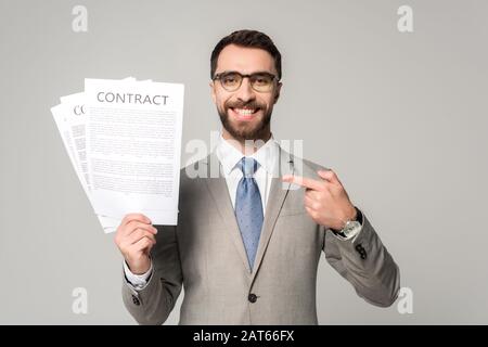 Lächelnder Geschäftsmann in Brille, der mit dem Finger auf Verträge zeigt, die auf grau isoliert sind Stockfoto