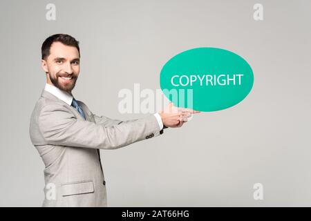 Gutaussehender Geschäftsmann, der Sprechblase mit Wort-Copyright hält und mit einer auf Grau isolierten Kamera lächelt Stockfoto
