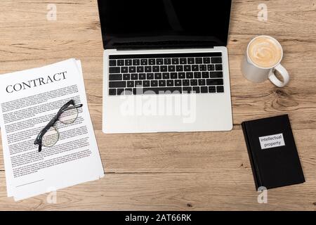 Draufsicht über Laptop, Vertrag, Brille, Tasse Kaffee und Buch über geistiges Eigentum auf Holzschreibtisch Stockfoto